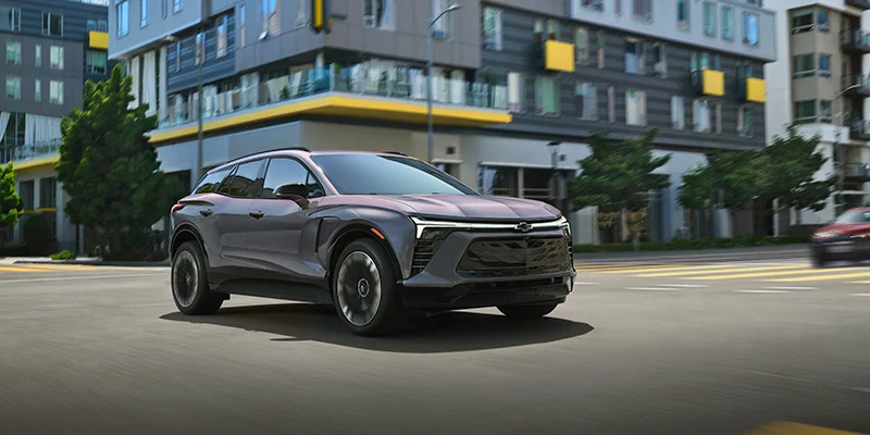 A sleek 2025 Chevy Blazer EV cruises through a downtown Roscommon, MI intersection on a sunny day, with modern high-rise apartments in the background.