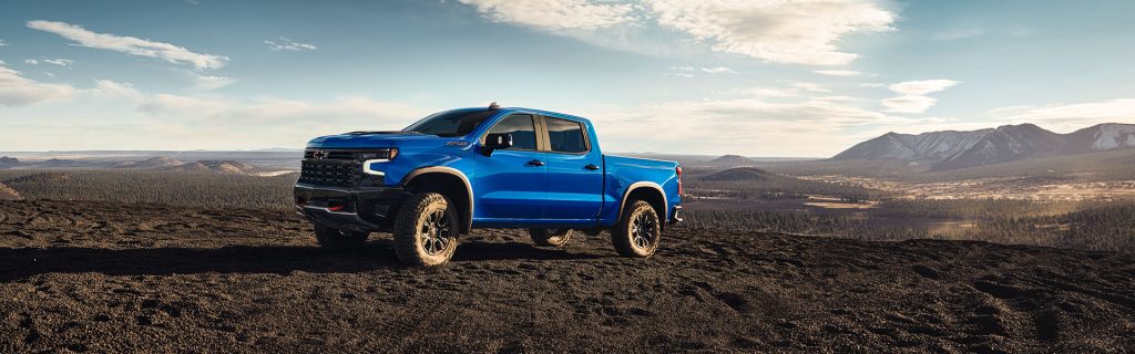 A blue 2025 Chevrolet Silverado 1500 driving through dark sand and soil in a desert-like terrain, with mountainous views in the background, near Don Nester Chevrolet in Roscommon, MI.