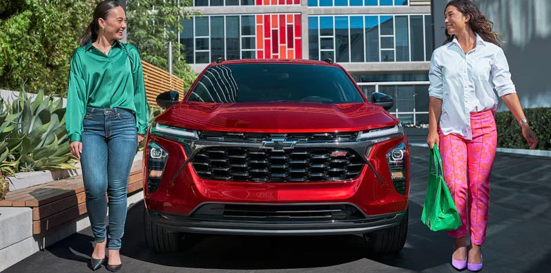 Two women walking around the front of a 2025 Chevrolet Traxs parked on the side of the road.