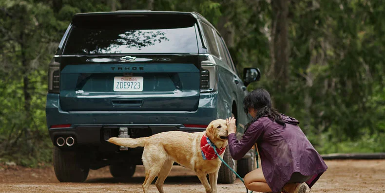 A woman petting her dog behind a parked blue 2026 Chevrolet Tahoe